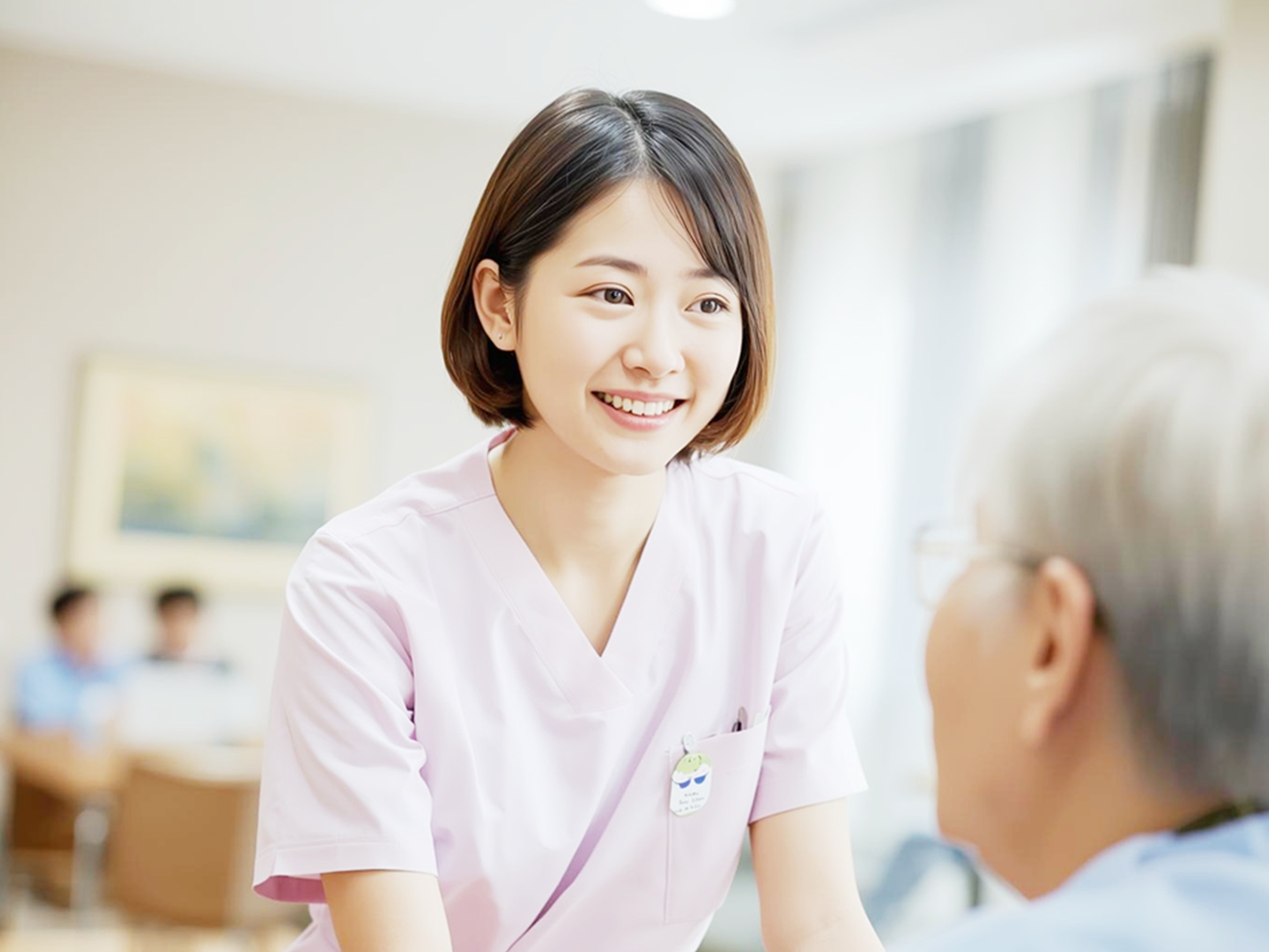 高齢患者に身をかがめて笑顔で語りかけるピンクのナース服の看護師